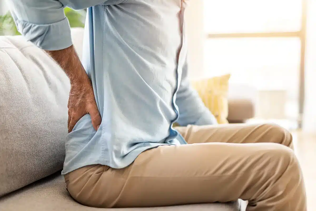 A person in pain sites on a couch and holds their lower back.